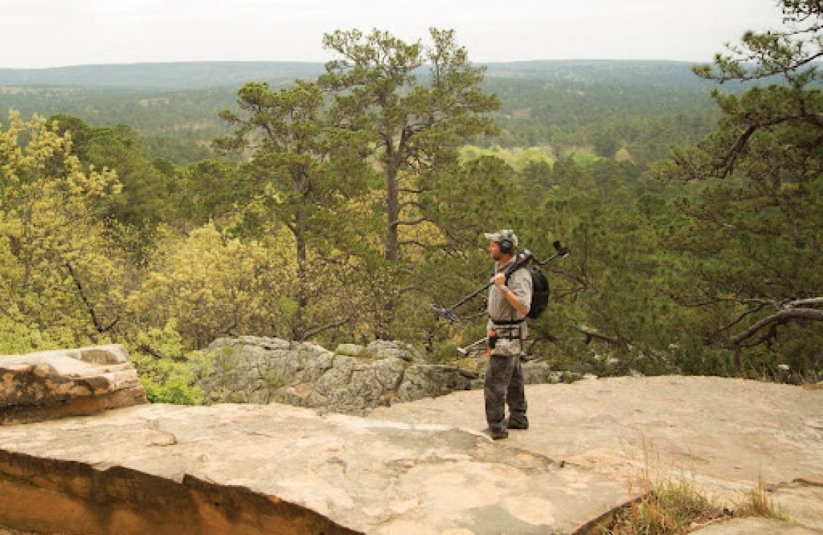 A detectorist on a rocky mountain overlooking a valley.